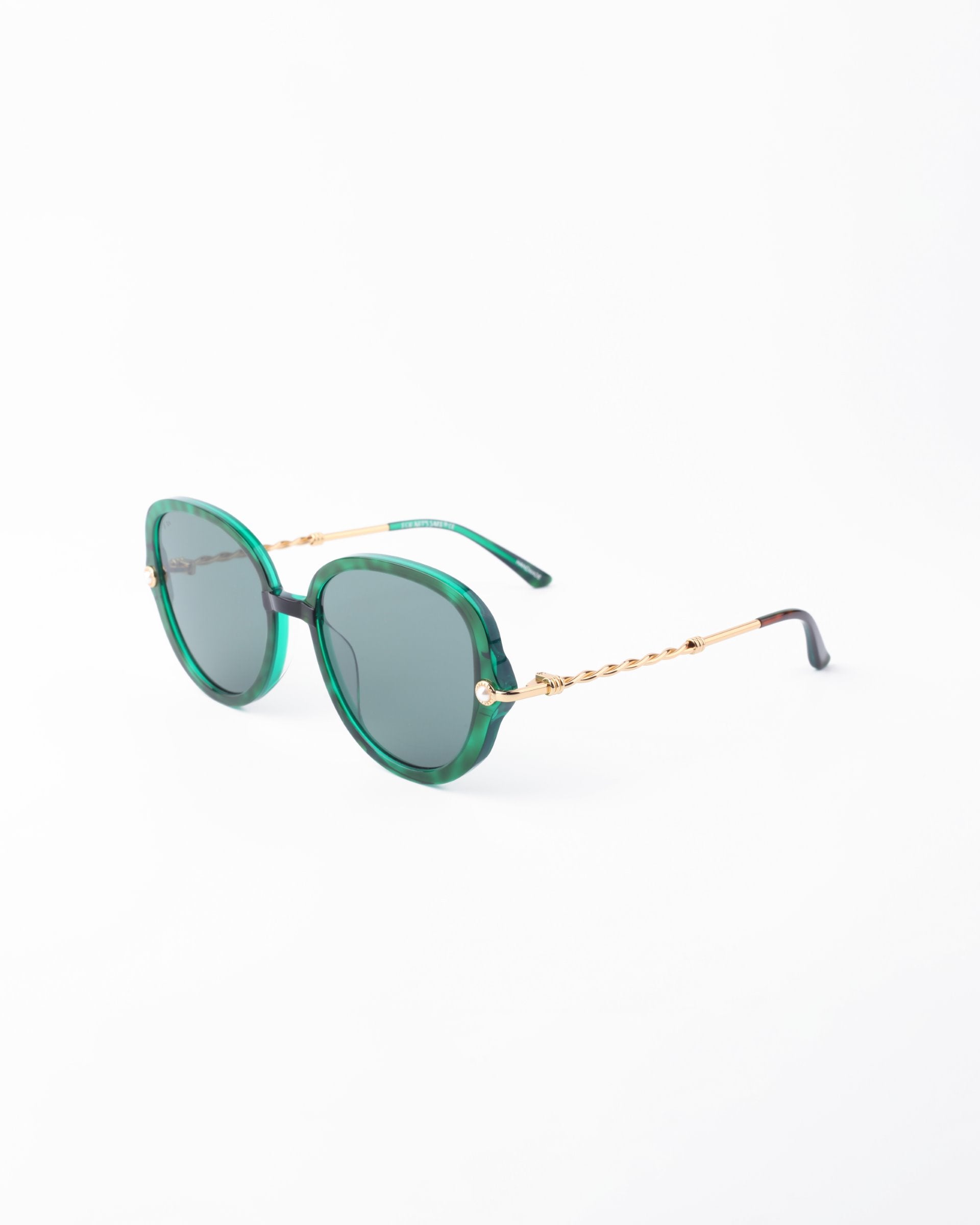 A pair of stylish For Art's Sake® Primrose sunglasses with green-tinted round lenses and handmade gold-plated metal arms. The frames are green with a glossy finish, made from plant-based acetate, and the twisted design on the arms adds a unique touch. The ends of the arms are black, and they feature an anti-reflective coating for enhanced comfort. Displayed on a white background.