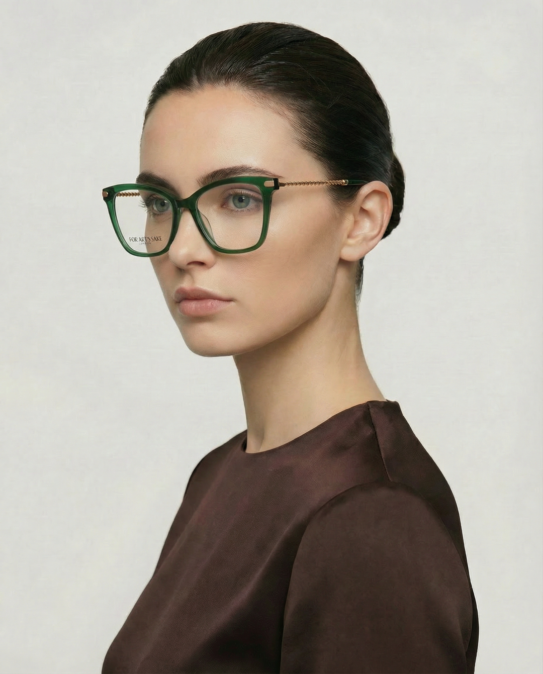 A woman with fair skin and dark hair in a neat updo wears For Art's Sake® Paris Two large green acetate eyewear and a brown top, looking slightly to the side against a plain, light background.