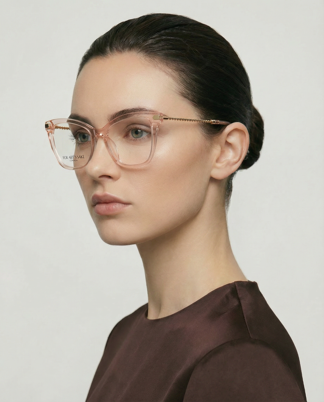 A woman with fair skin and dark hair in a low bun wears For Art's Sake® Paris Two—large, transparent acetate glasses—with a dark brown top, posing in profile against a plain, light background.