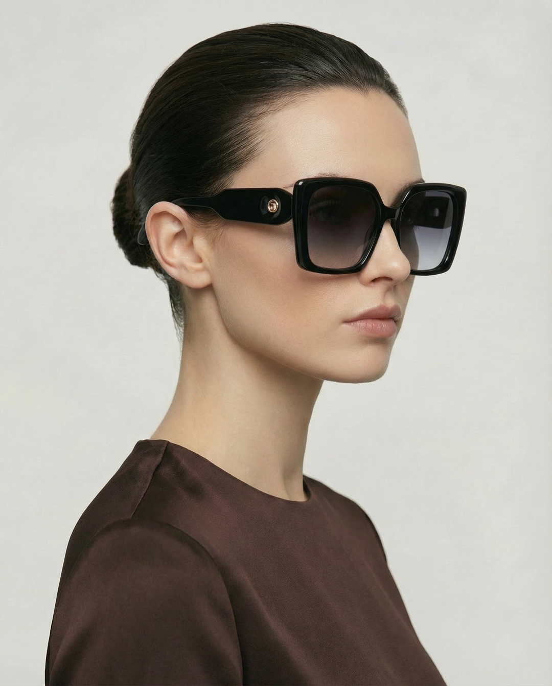 A woman with dark hair in a low bun wears For Art's Sake® Eos oversized soft-square sunglasses and a brown top, posing against a plain light background.