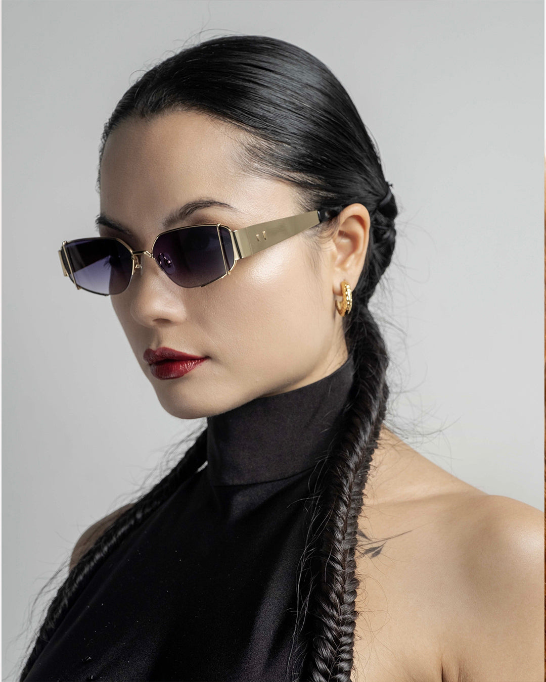 A woman with sleek, braided hair wears For Art's Sake® Talia gold-plated, full metal frame sunglasses, gold hoops, a sleeveless black top, and red lipstick while posing against a plain light background.