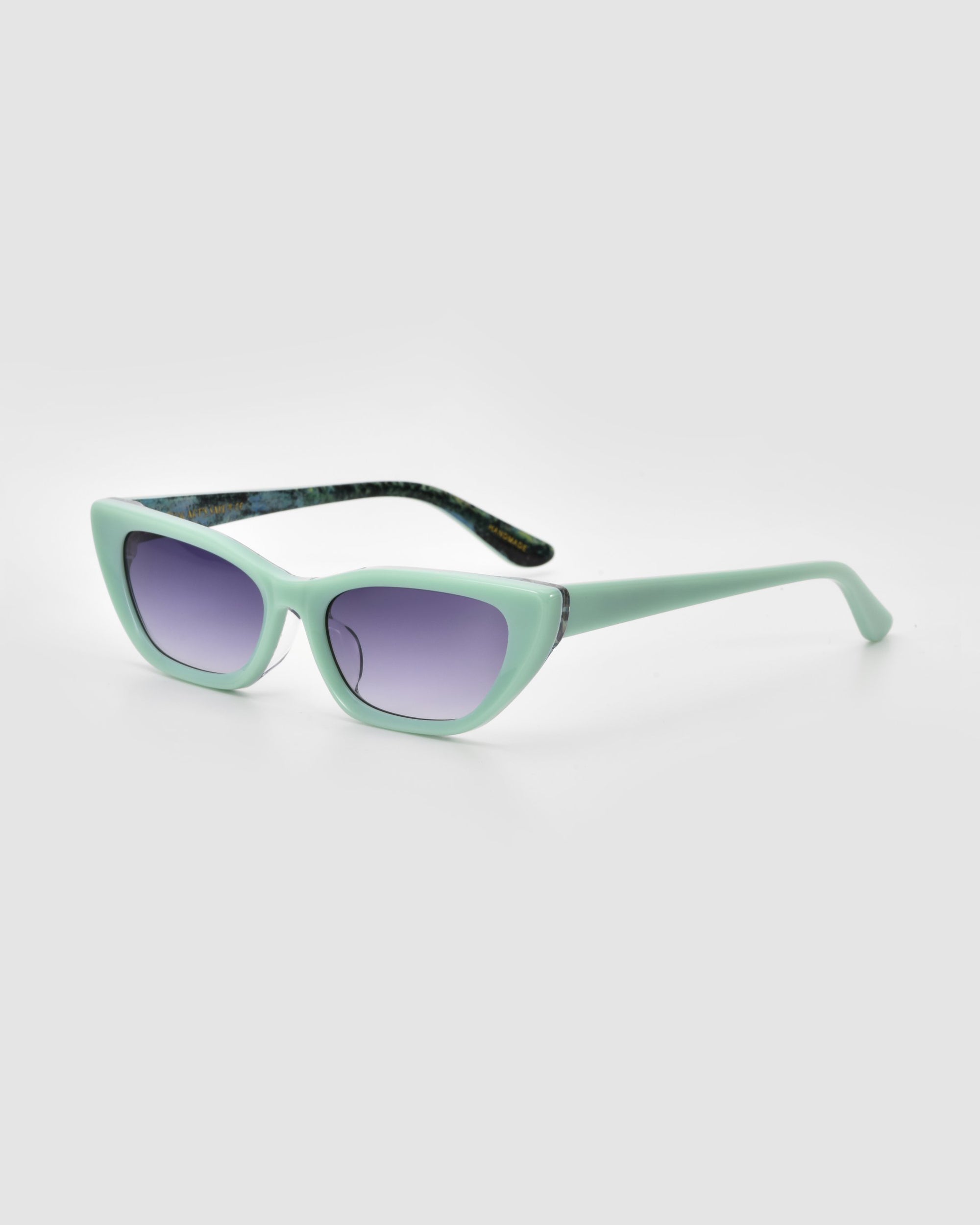 Claude Monet cat-eye sunglasses by For Art's Sake® feature a mint green frame, purple-tinted lenses, and dark patterned inner arms. Inspired by Monet’s elegance, these limited edition shades are shown on a light gray background.