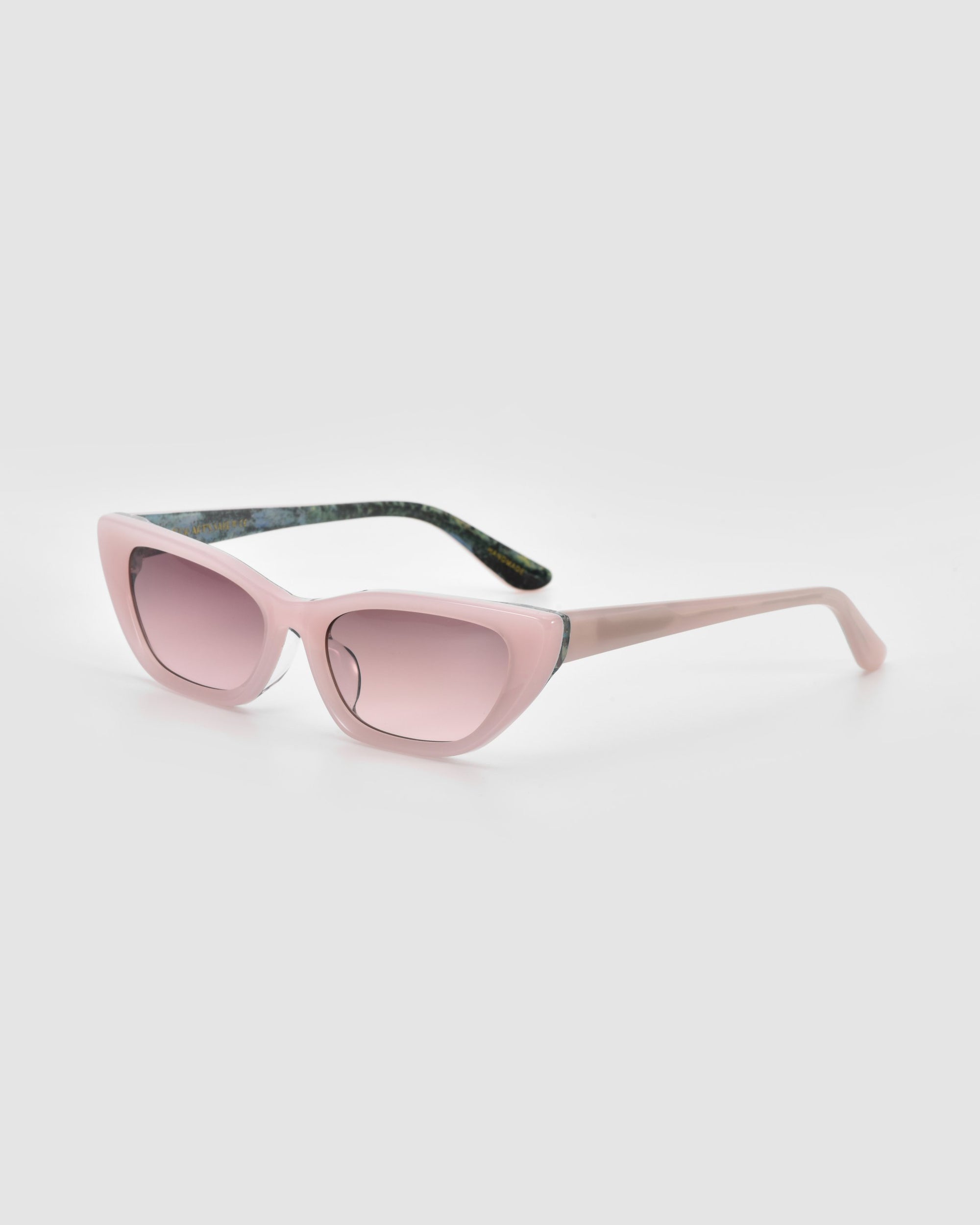 The Claude Monet by For Art's Sake® are limited edition cat-eye sunglasses with light pink frames, matching tinted lenses, and dark green patterned arms, shown on a light gray background.