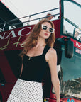 A woman with long red hair wears For Art's Sake® Dusk chunky cat-eye sunglasses, a black sleeveless top, and a white polka dot skirt while standing in front of a red open-top tour bus on a sunny day.
