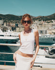 Wearing For Art's Sake® Bloom oversized acetate sunglasses, a woman in a white tank top and skirt stands by a marina with yachts and a coastal city with hills in the background on a sunny day.