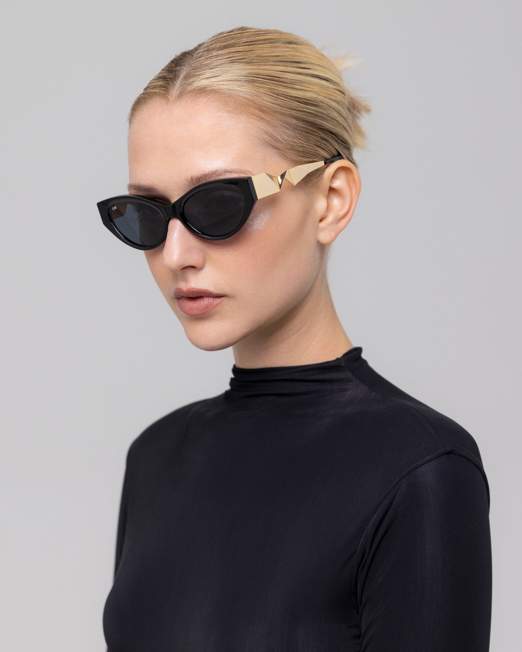 A woman with blonde hair in a bun wears For Art's Sake® Unfold black cat-eye sunglasses with gold accents and a black turtleneck, standing against a light gray background, her look elevated by the sculptural frames.