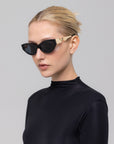 A woman with blonde hair in a bun wears For Art's Sake® Unfold black cat-eye sunglasses with gold accents and a black turtleneck, standing against a light gray background, her look elevated by the sculptural frames.