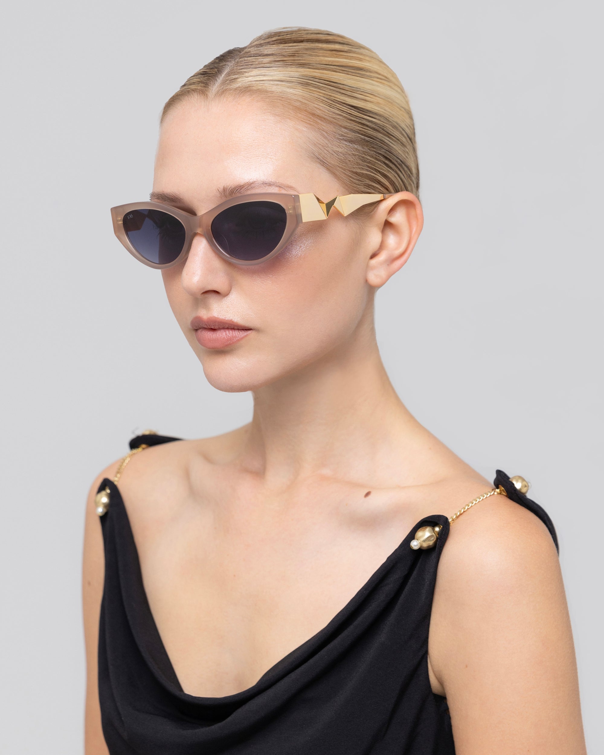 A woman with blonde hair in a sleek bun wears Unfold sculptural beige sunglasses by For Art's Sake® and a black sleeveless top with gold chain straps, looking off to the side against a plain light gray background.