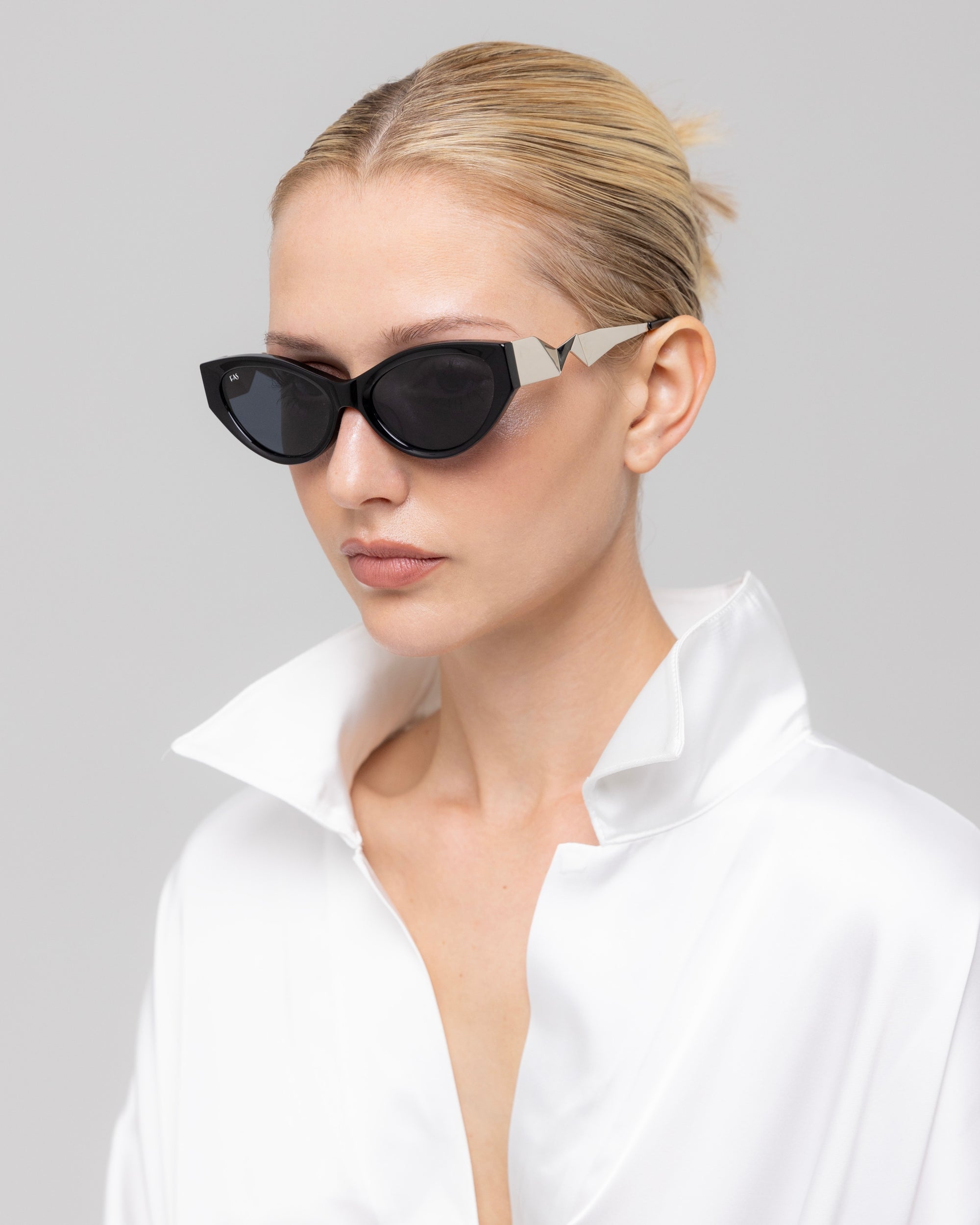 A person with light hair tied back wears For Art's Sake® Unfold black sculptural cat-eye sunglasses and a shiny white collared shirt, looking to the side against a plain, light gray background.