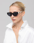 A person with light hair tied back wears For Art's Sake® Unfold black sculptural cat-eye sunglasses and a shiny white collared shirt, looking to the side against a plain, light gray background.