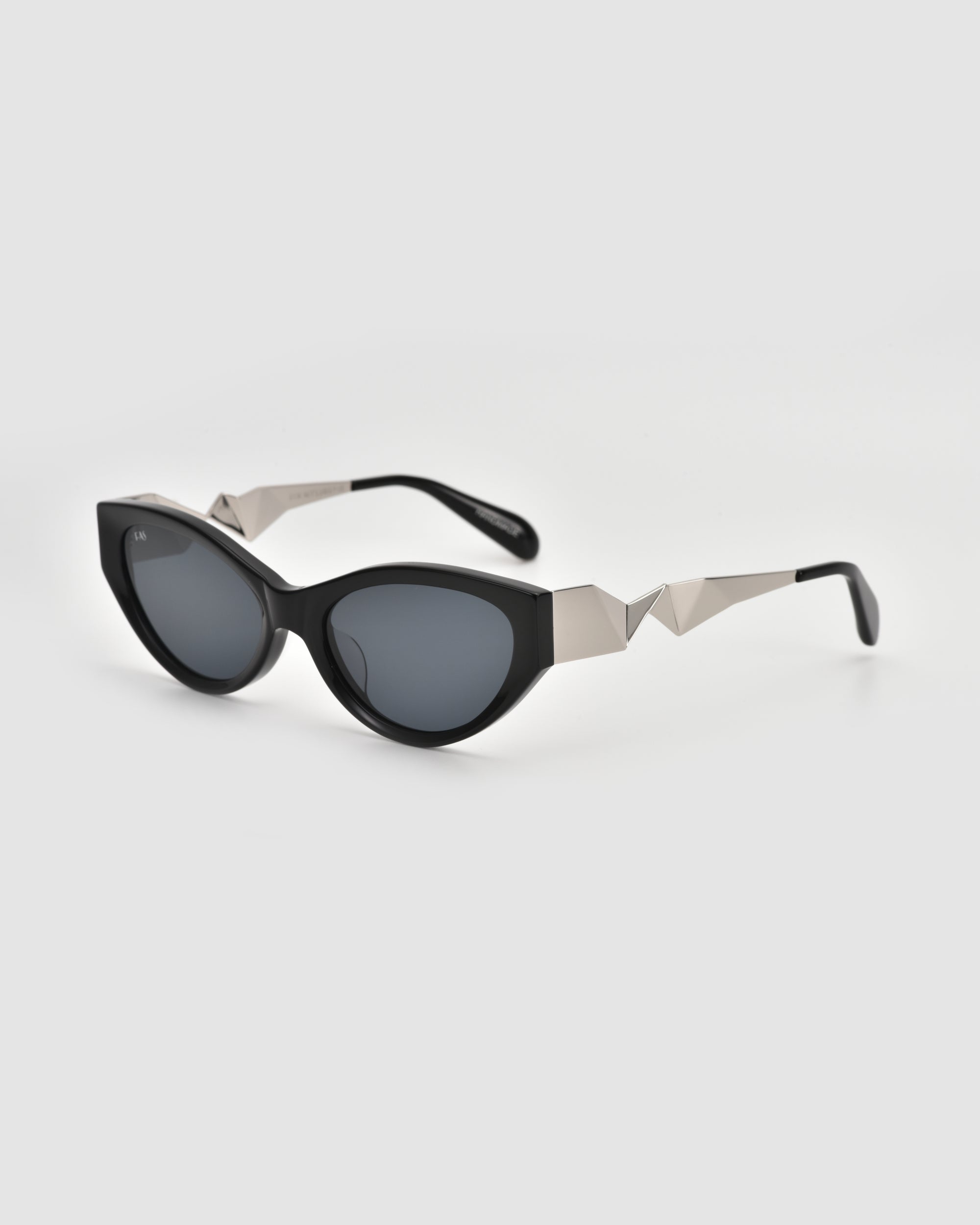 For Art’s Sake® Unfold black oval cat-eye sunglasses feature dark lenses and striking angular silver arms, making a bold geometric statement on a plain light background.