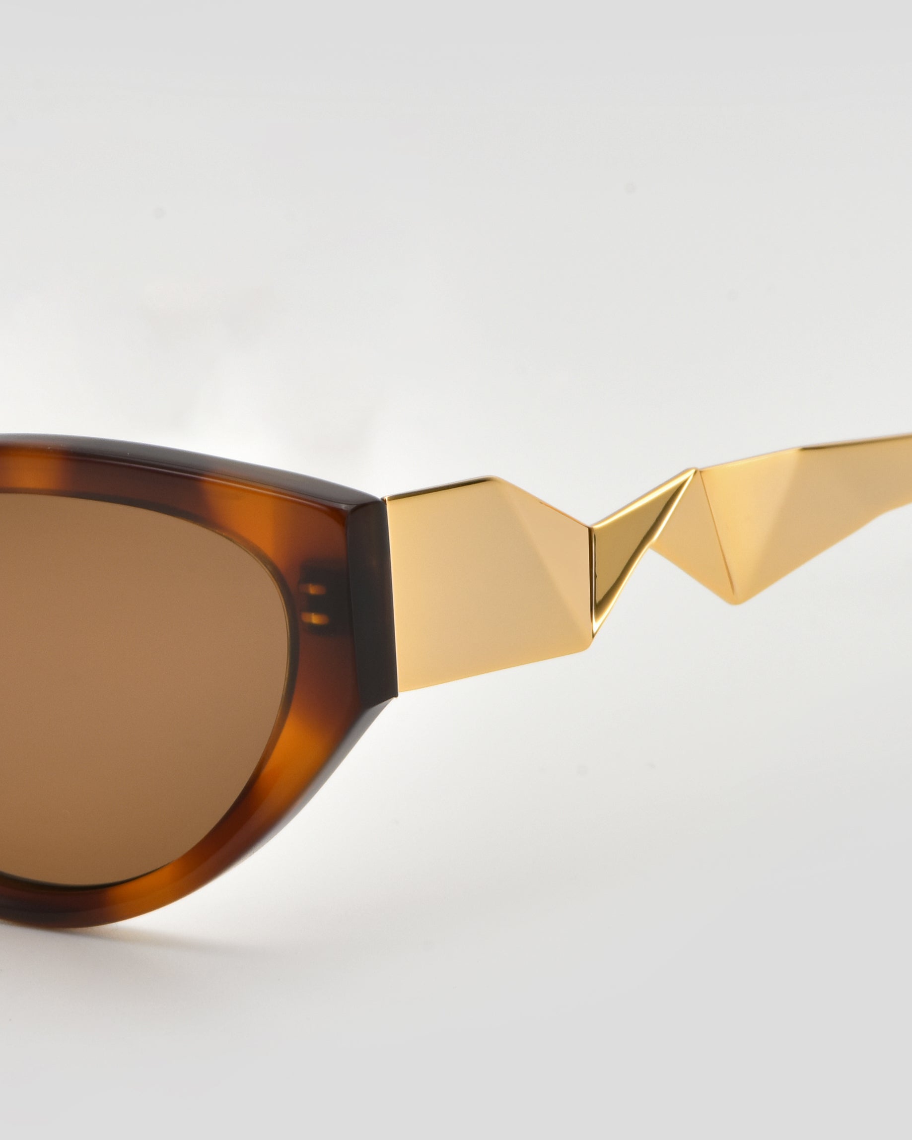 Close-up of For Art’s Sake® Unfold sunglasses featuring brown lenses and ornate gold geometric details on pleated temples, set against a plain light background.