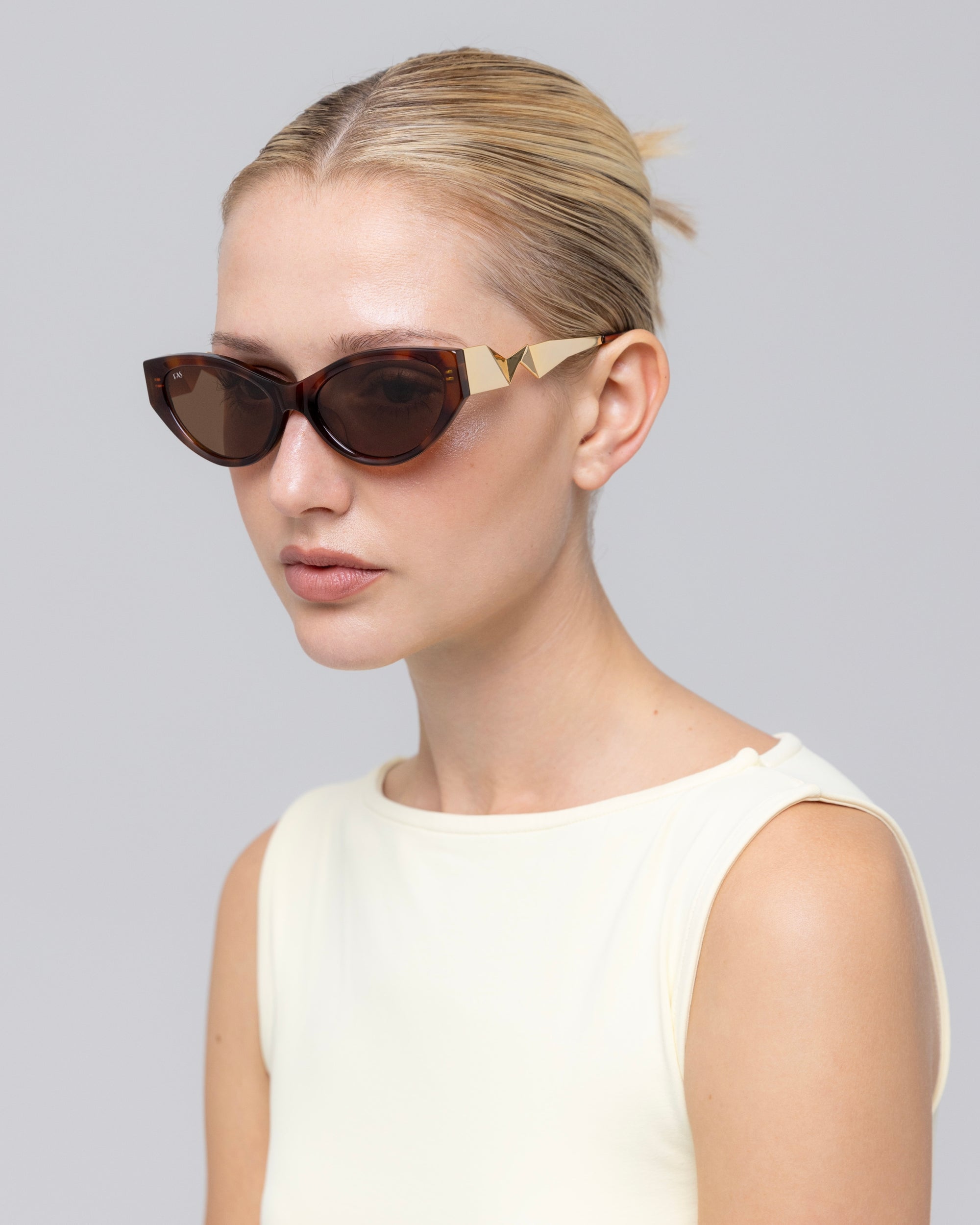 A woman with blonde hair in a neat bun wears the Unfold geometric cat-eye sunglasses by For Art's Sake®, featuring gold accents, paired with a sleeveless light top against a plain light gray background.