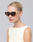 A woman with blonde hair in a neat bun wears the Unfold geometric cat-eye sunglasses by For Art's Sake®, featuring gold accents, paired with a sleeveless light top against a plain light gray background.
