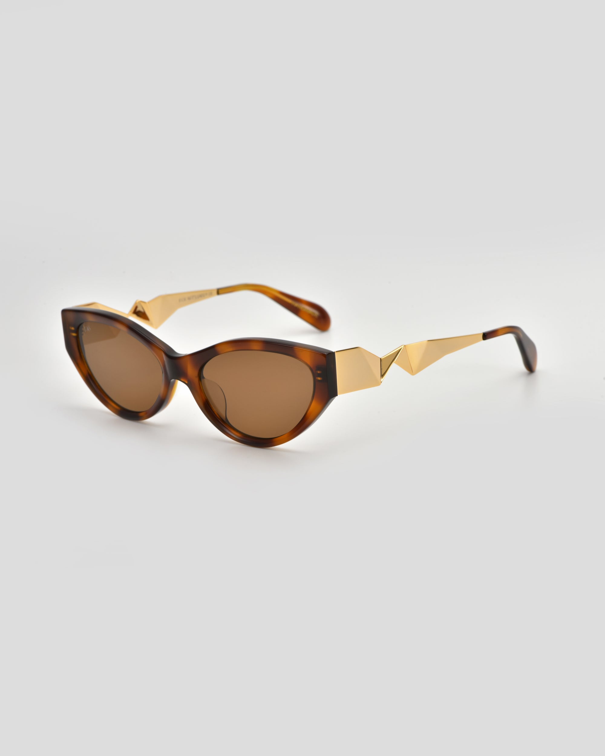 For Art's Sake® Unfold sunglasses: tortoiseshell cat-eye frames with brown lenses and gold geometric arm detailing, showcased as striking geometric eyewear on a light gray background.