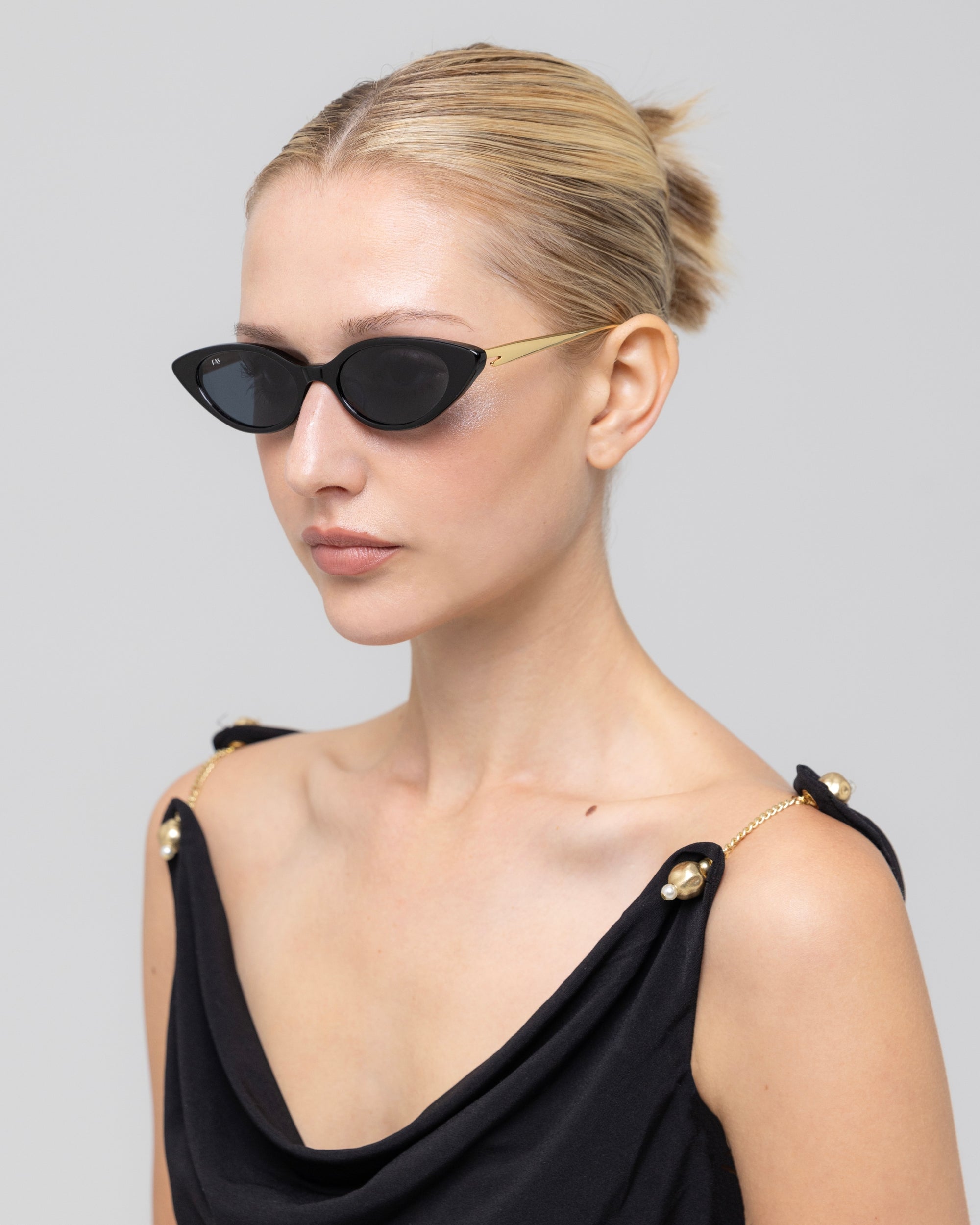 A woman with blonde hair pulled back wears For Art's Sake® Slash sculptural cat-eye sunglasses and a black sleeveless top with gold accents, glancing to the side against a plain light background.
