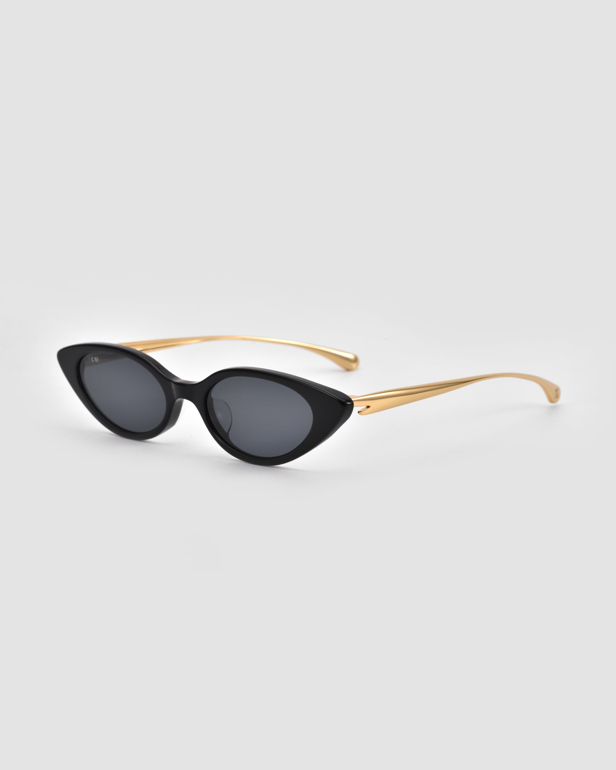 The Slash sunglasses by For Art's Sake® feature black cat-eye frames, dark lenses, and sleek gold arms, capturing the bold allure of sculptural eyewear against a light gray background.