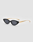 The Slash sunglasses by For Art's Sake® feature black cat-eye frames, dark lenses, and sleek gold arms, capturing the bold allure of sculptural eyewear against a light gray background.
