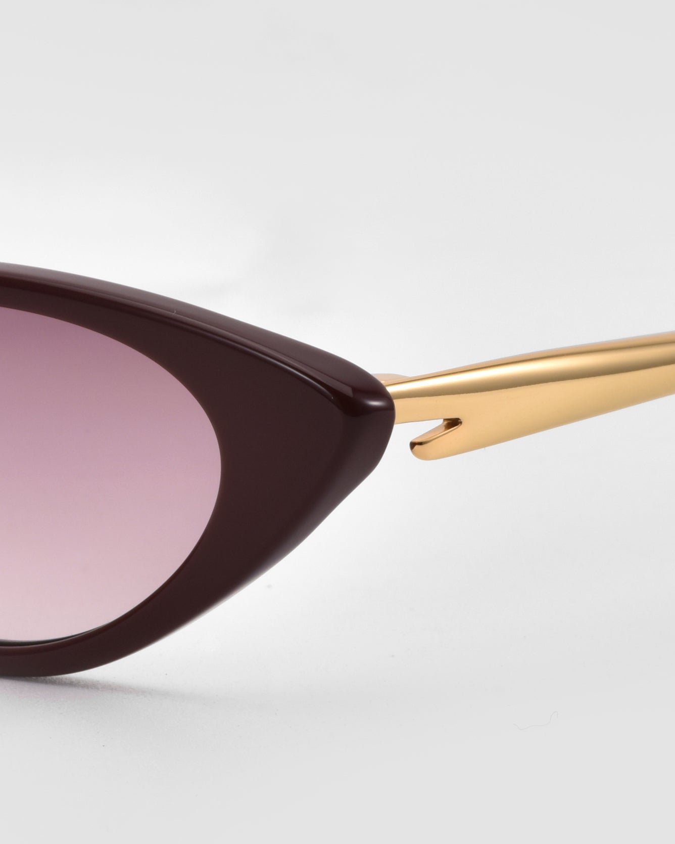 Close-up of For Art&#39;s Sake® Slash cat-eye sunglasses with a dark burgundy frame, pink-tinted lens, and gold metal arm, highlighting sculpted details against a light gray background.