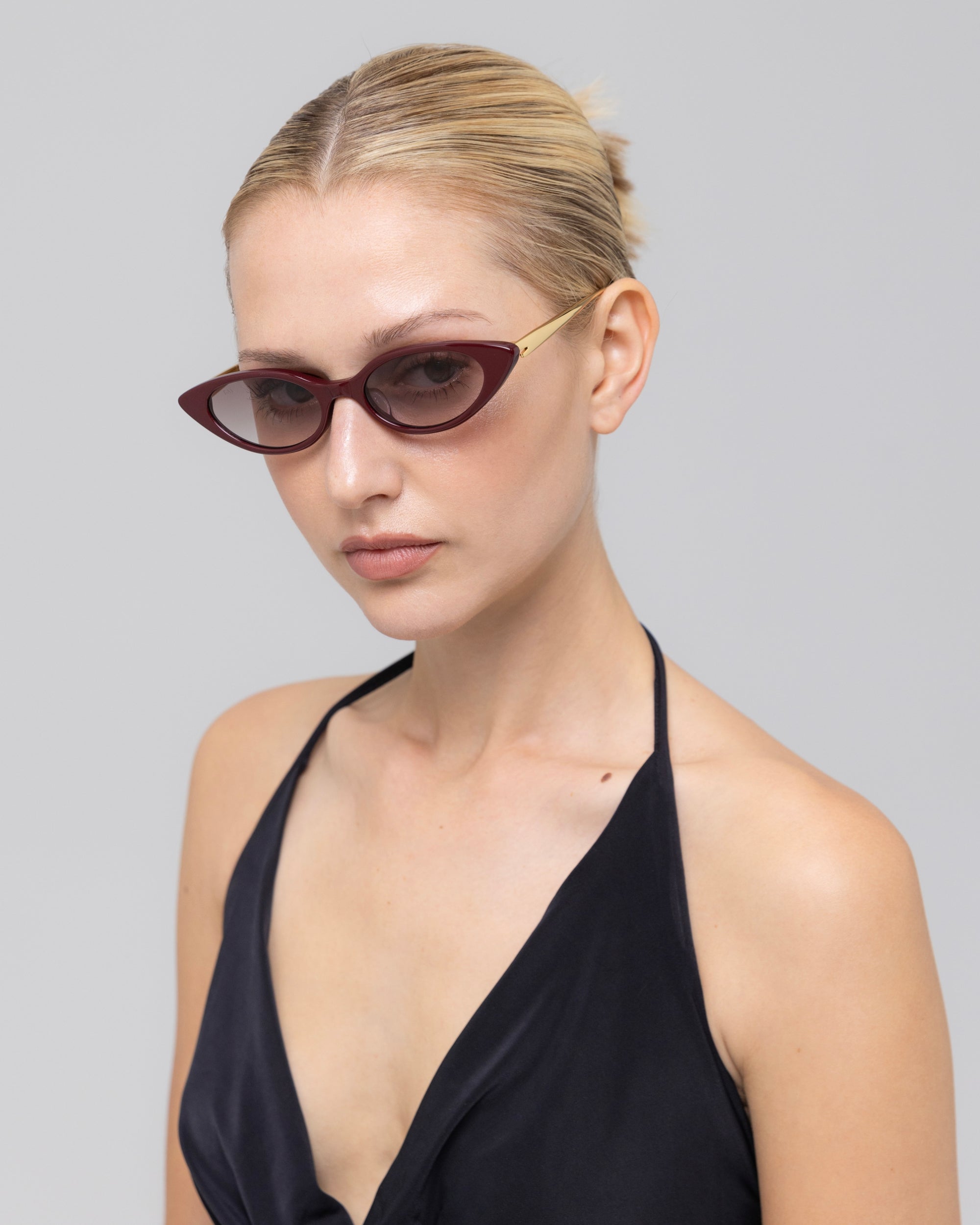 A woman with blonde hair in a sleek bun wears For Art's Sake® Slash sculptural cat-eye sunglasses with dark lenses and a black halter top, posing against a plain light gray background.