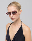 A woman with blonde hair in a sleek bun wears For Art's Sake® Slash sculptural cat-eye sunglasses with dark lenses and a black halter top, posing against a plain light gray background.