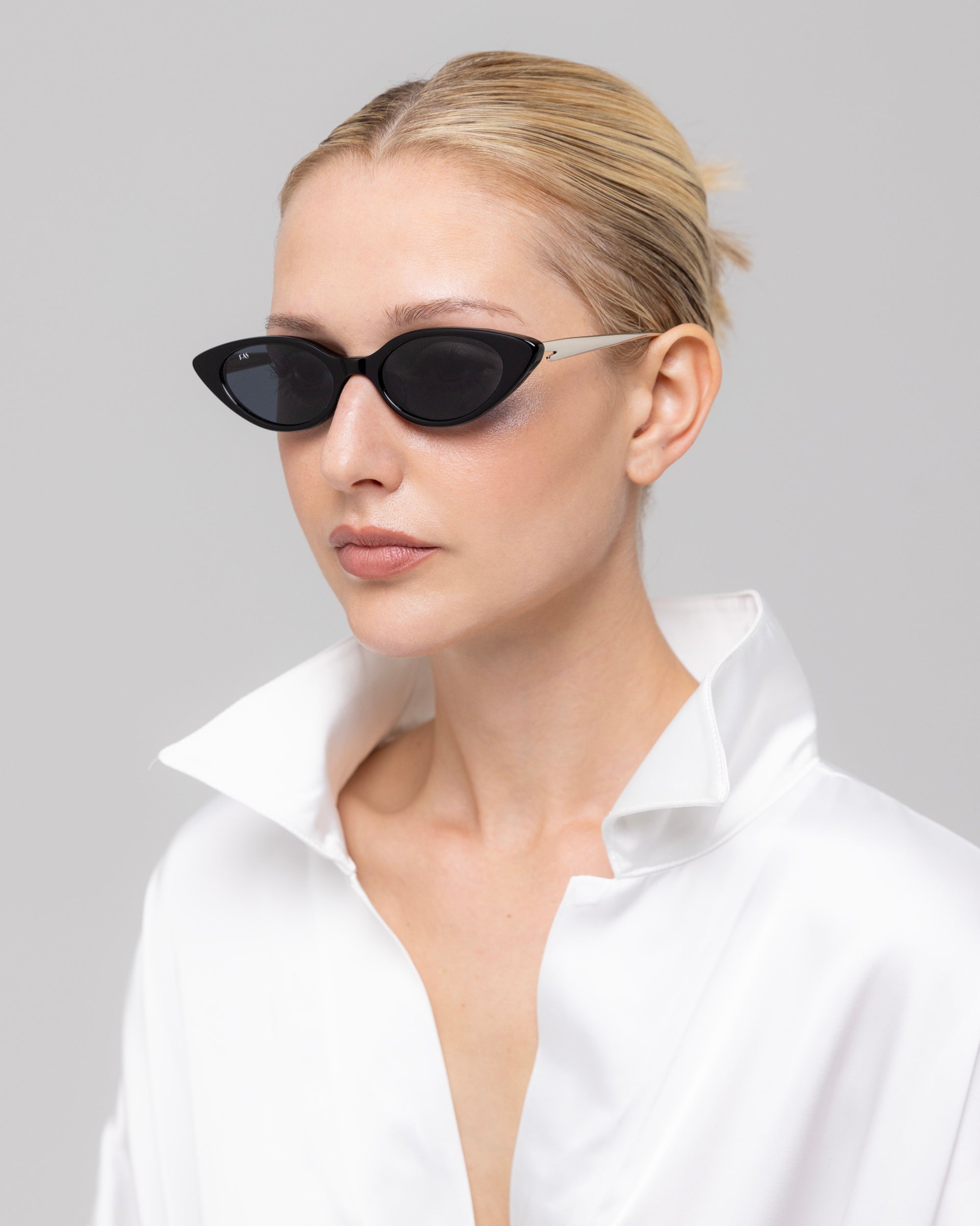 A woman with blond hair in a sleek bun models For Art's Sake® Slash black oval sunglasses and a white satin shirt with a wide open collar, posing against a plain light gray background.