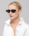 A woman with blond hair in a sleek bun models For Art's Sake® Slash black oval sunglasses and a white satin shirt with a wide open collar, posing against a plain light gray background.