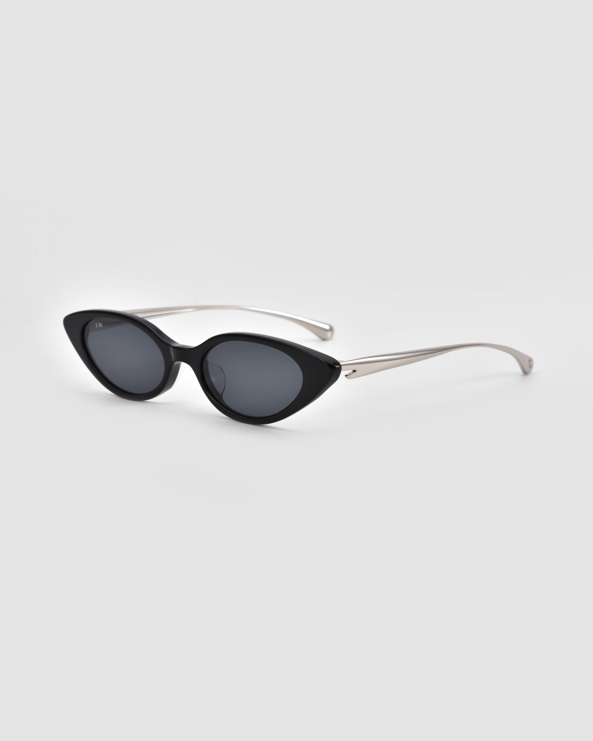 The Slash sunglasses by For Art's Sake® feature a black cat-eye design, dark lenses, and silver metal arms, showcased against a light gray background.