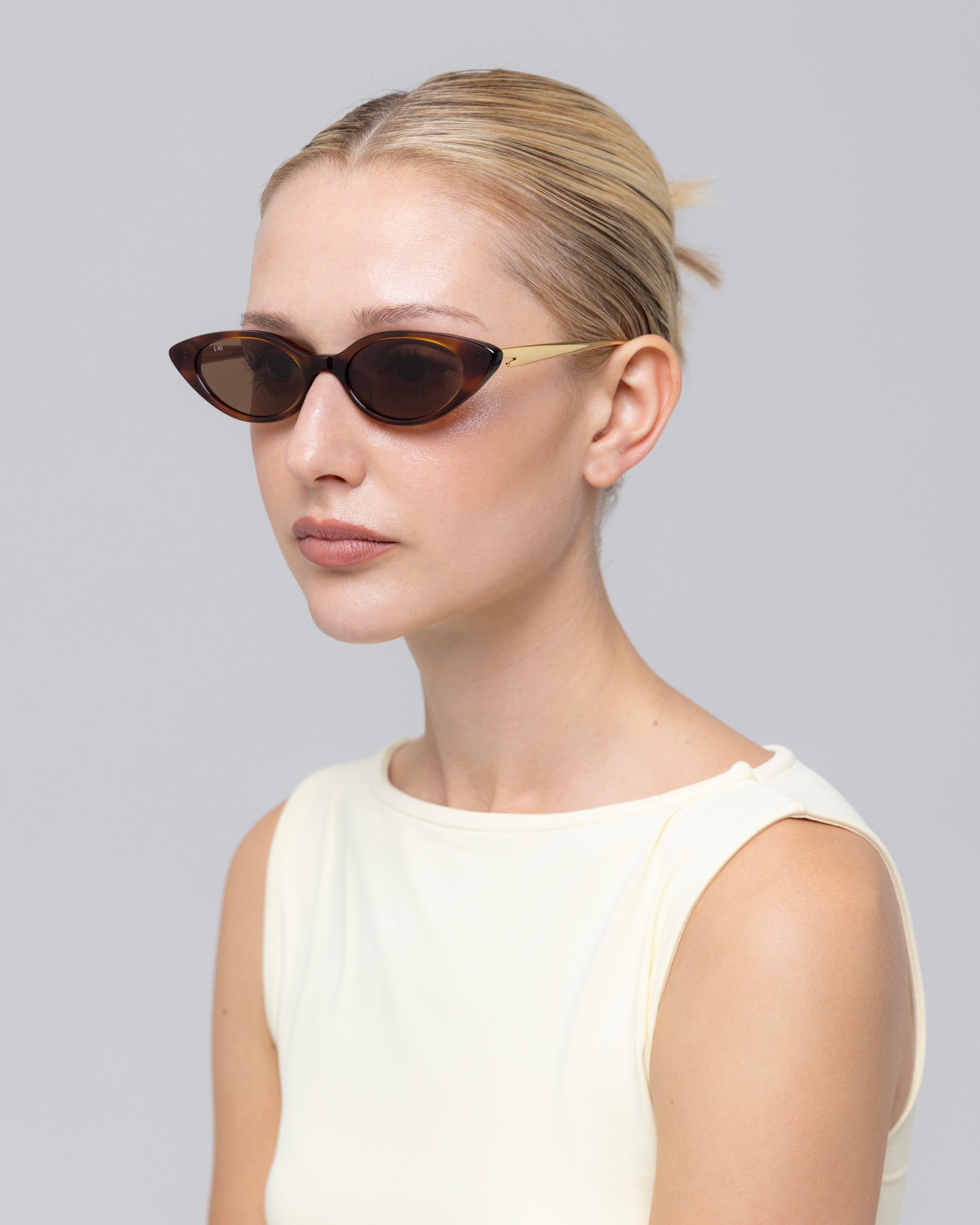 A woman with a low blonde bun wears For Art's Sake® Slash sculptural cat-eye sunglasses and a sleeveless off-white top, posing against a plain light grey background.