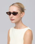 A woman with a low blonde bun wears For Art's Sake® Slash sculptural cat-eye sunglasses and a sleeveless off-white top, posing against a plain light grey background.