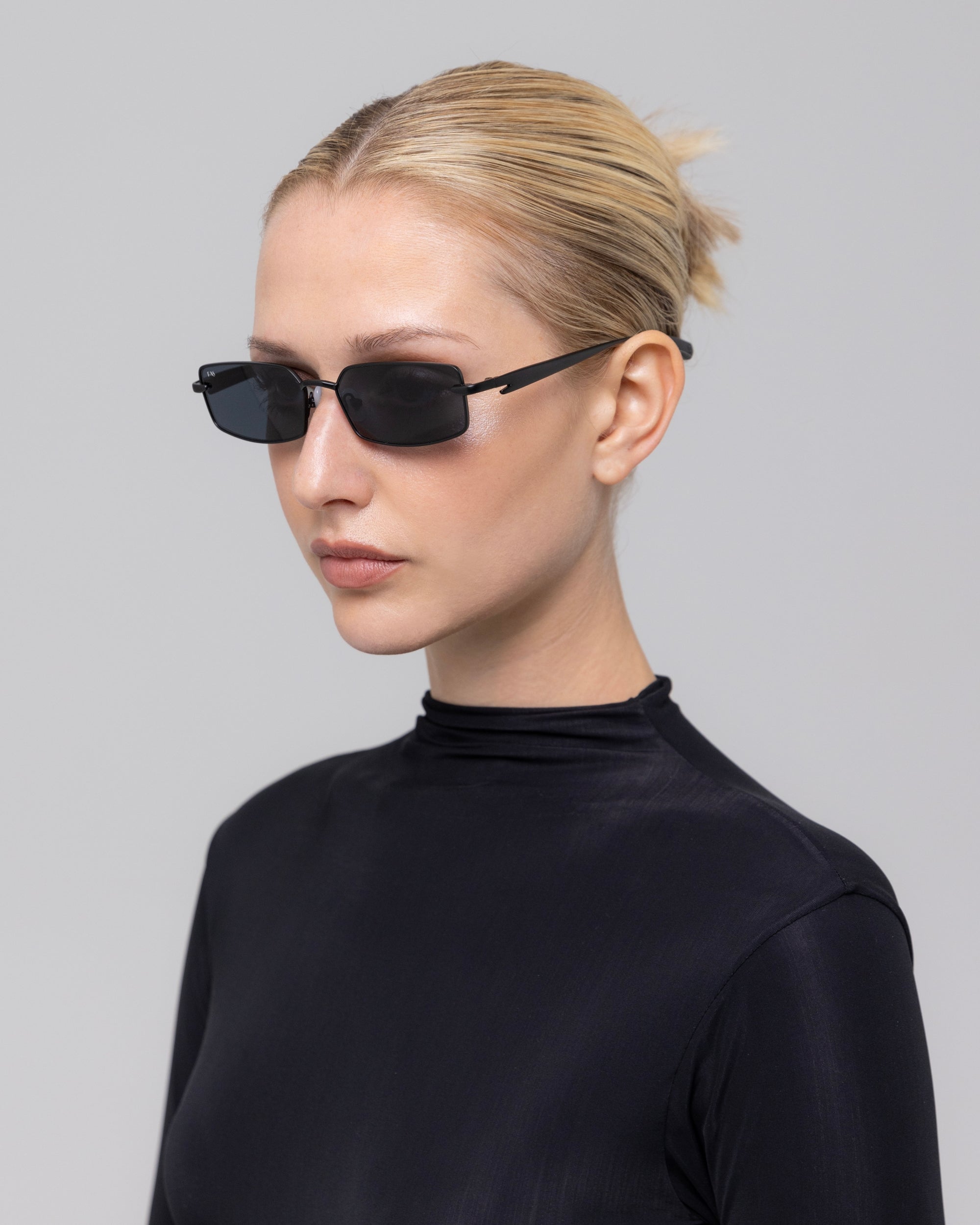 A woman with blonde hair tied back, wearing For Art's Sake® Lucio sculpted eyewear featuring a 3D swoosh temple and a black high-neck top, stands against a plain light gray background.