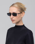 A woman with blonde hair tied back, wearing For Art's Sake® Lucio sculpted eyewear featuring a 3D swoosh temple and a black high-neck top, stands against a plain light gray background.
