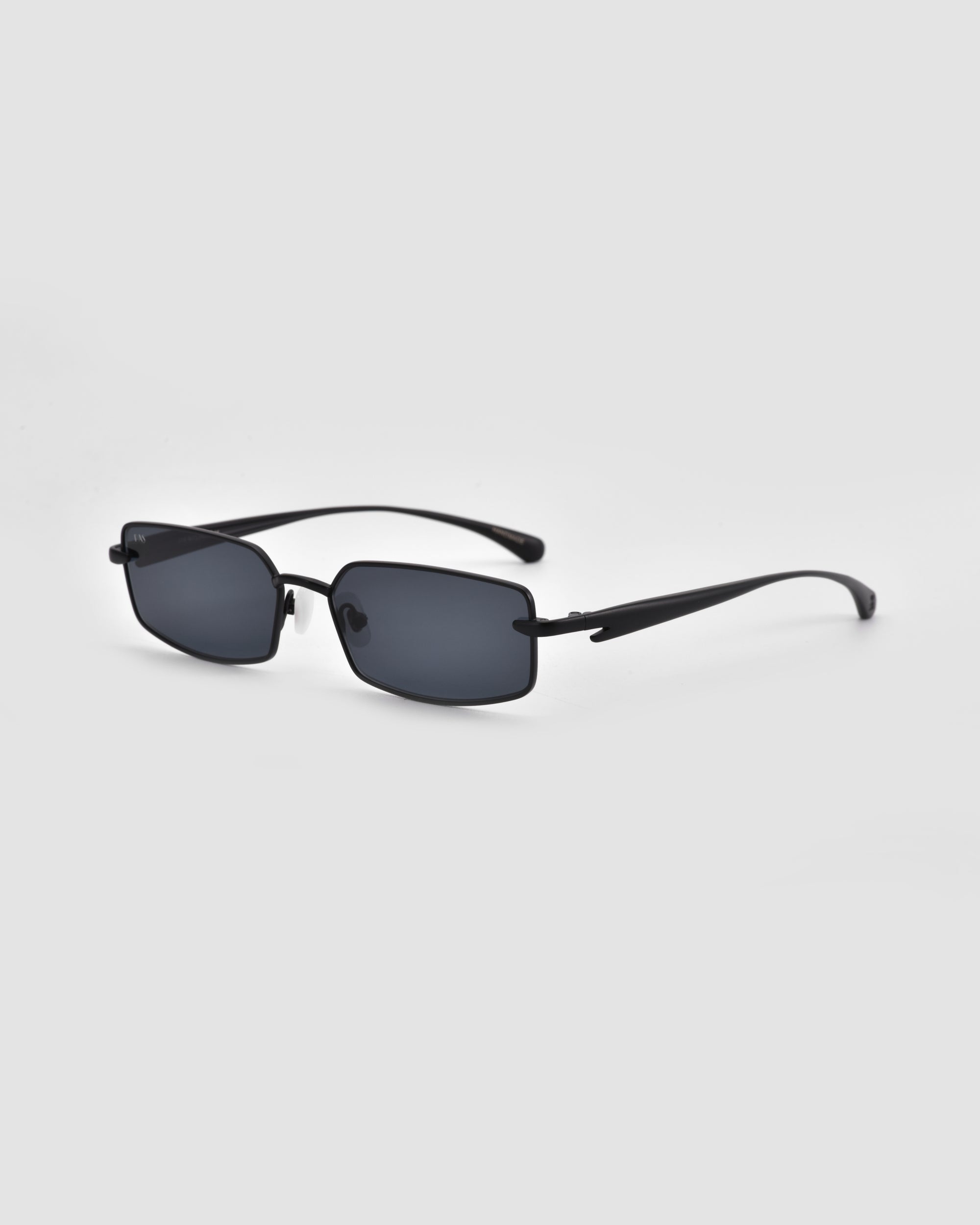 The Lucio by For Art's Sake® features black rectangular sunglasses with thin frames, dark lenses, and sculpted details, showcased on a plain light gray background.