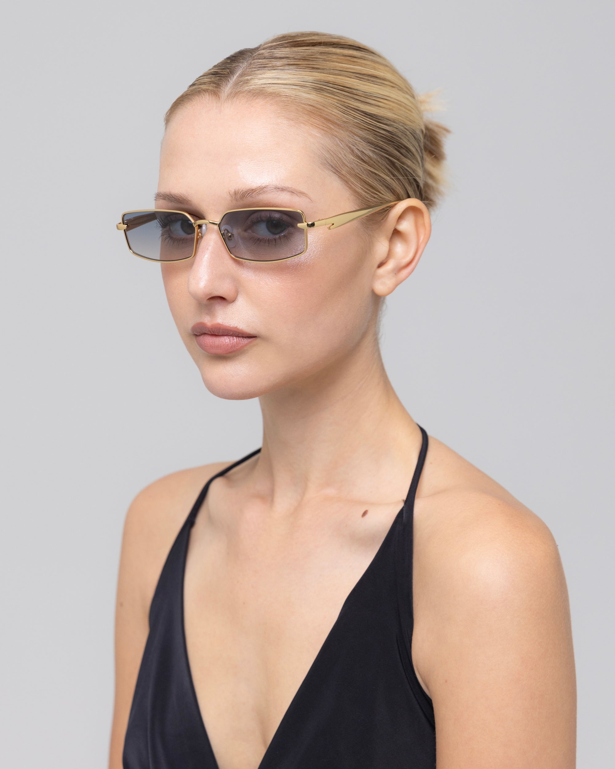 A blonde woman with her hair tied back wears For Art's Sake® Lucio sculpted rectangular eyewear and a black halter-neck top, gazing slightly to the side against a plain gray backdrop.