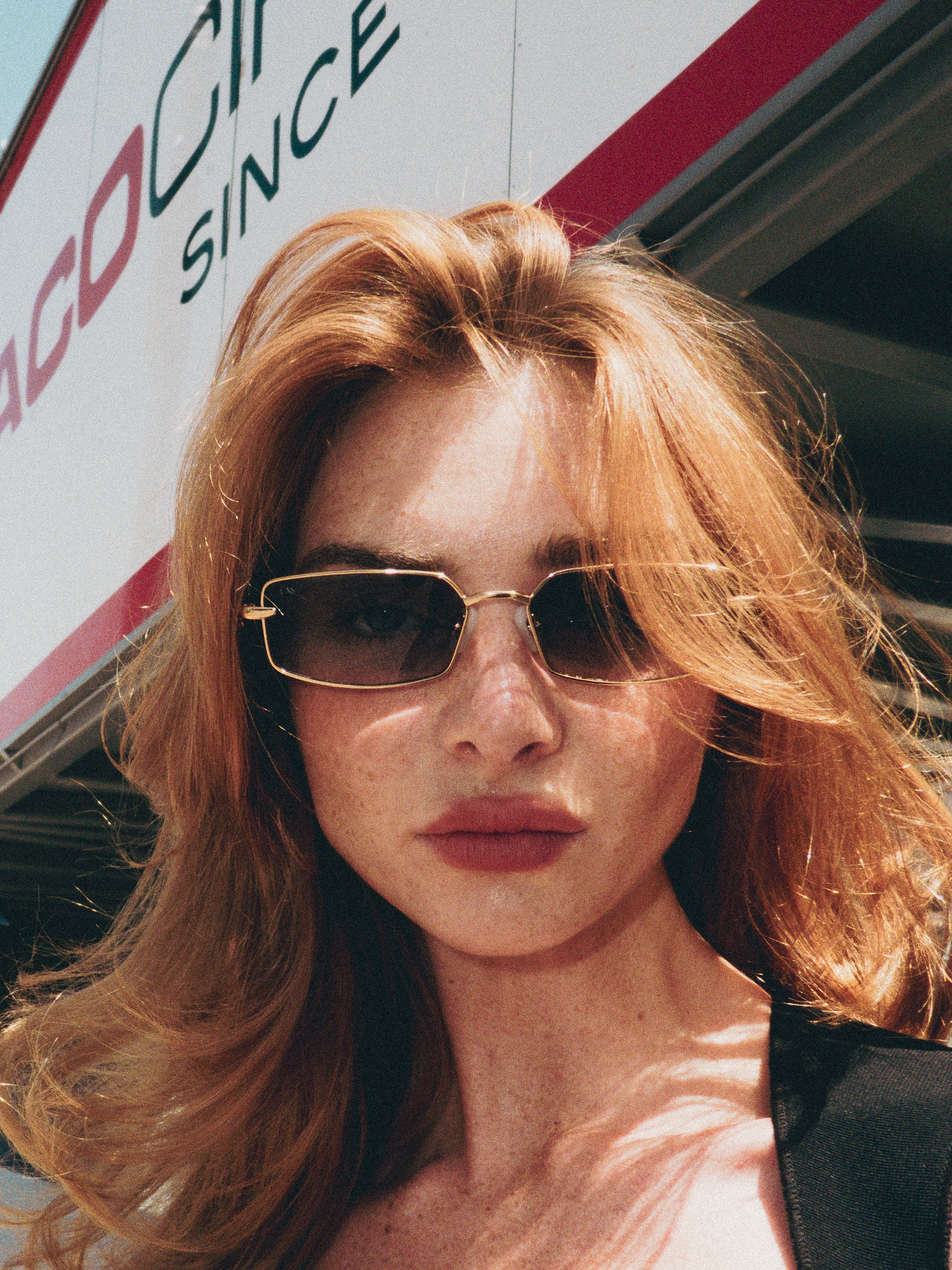 A woman with long red hair wears For Art’s Sake® Lucio sunglasses outside, with a building and an "ACO" sign in the background.