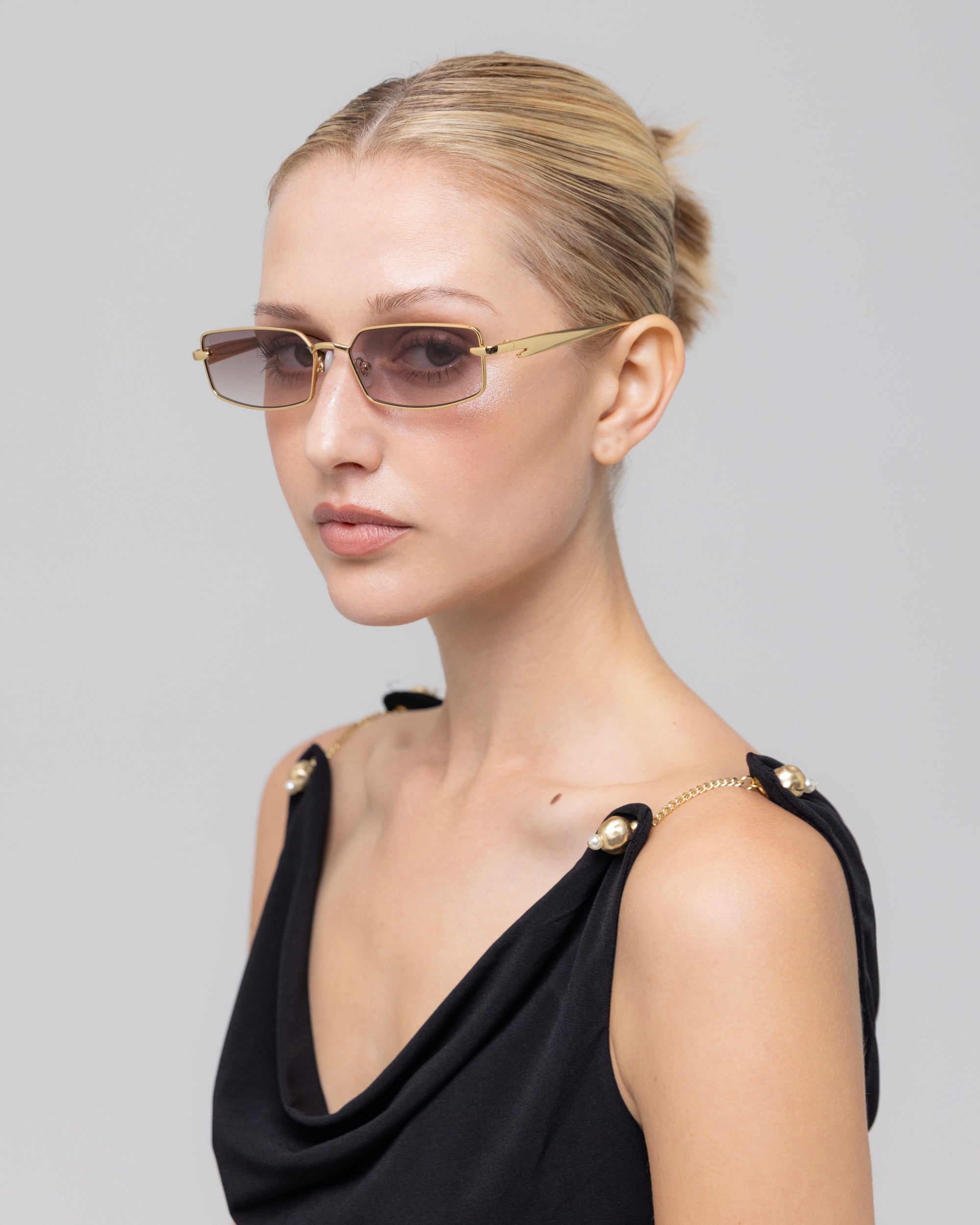 A woman with blonde hair in a bun models For Art's Sake® Lucio rectangular sunglasses with gold button accents, paired with a black sleeveless top, against a plain light gray background.