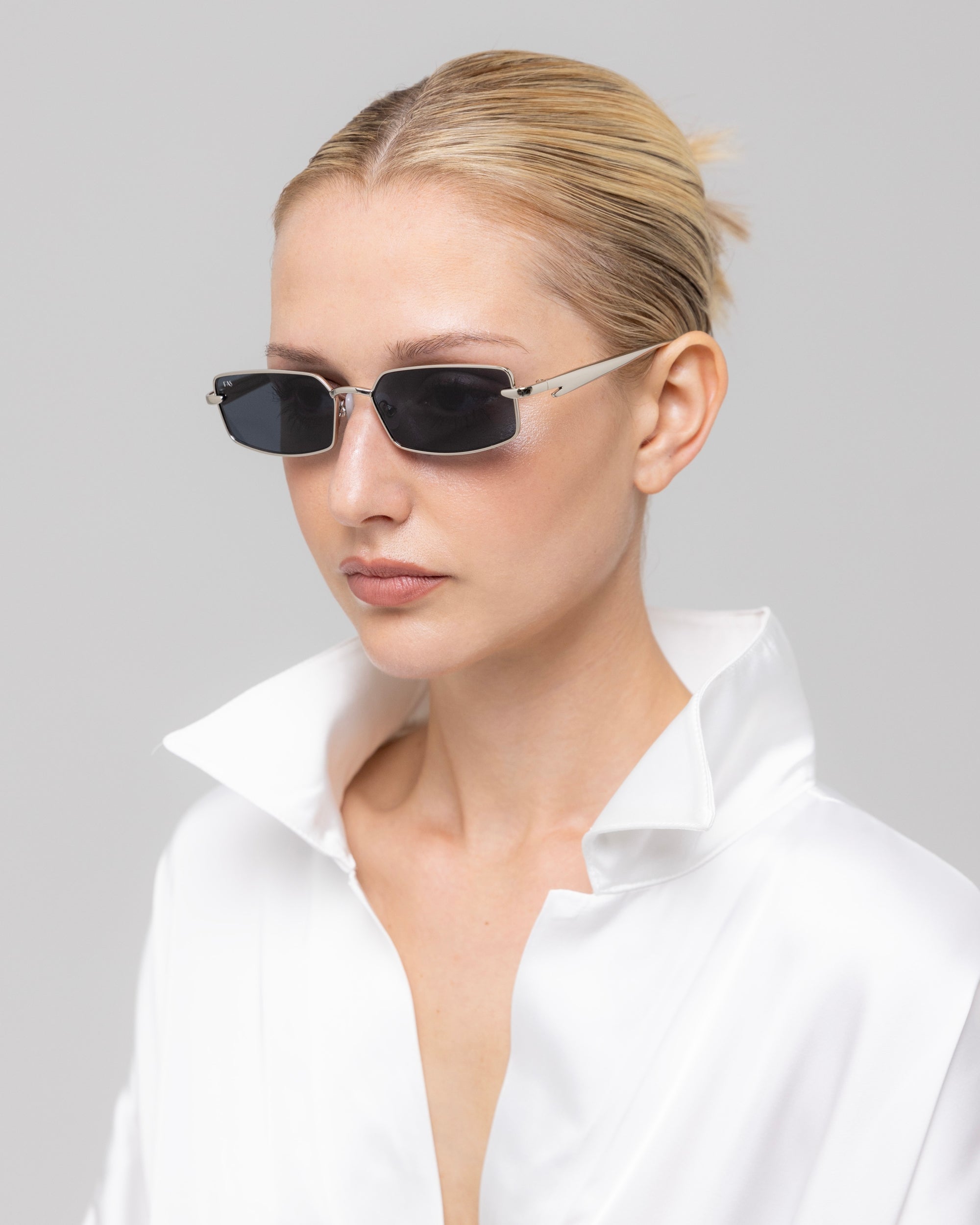 A woman with blonde hair in a sleek bun wears For Art's Sake® Lucio narrow black cat-eye sunglasses and a shiny white collared blouse, looking to the side against a plain light gray background.