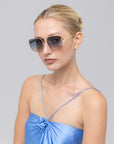 A woman with blonde hair in a sleek bun wears For Art's Sake® Edge sunglasses, featuring a bold rectangular silhouette, and a shiny light blue spaghetti strap top while posing against a plain light gray background.