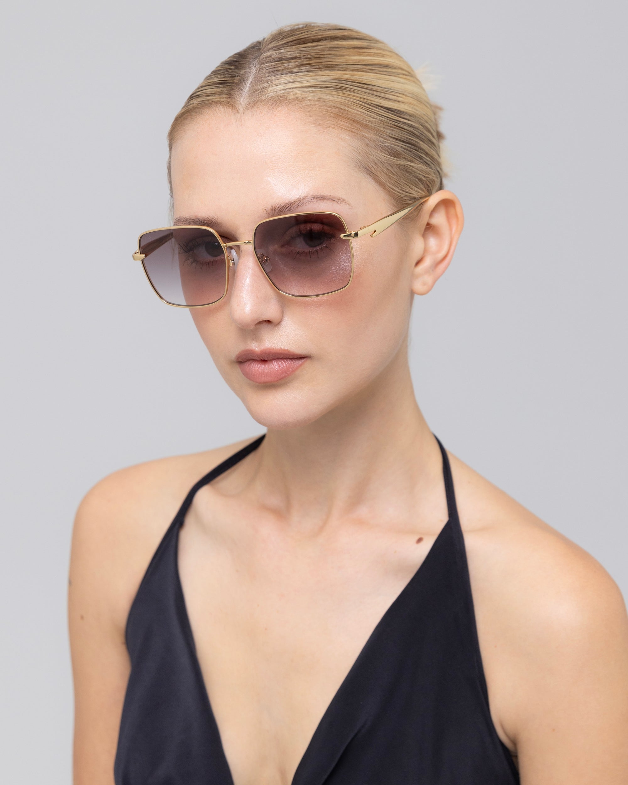 A woman with blonde hair tied back wears For Art's Sake® Edge sunglasses, featuring a rectangular silhouette and gold frames. She pairs them with a black halter top and poses against a plain light gray background.