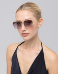 A woman with blonde hair tied back wears For Art's Sake® Edge sunglasses, featuring a rectangular silhouette and gold frames. She pairs them with a black halter top and poses against a plain light gray background.