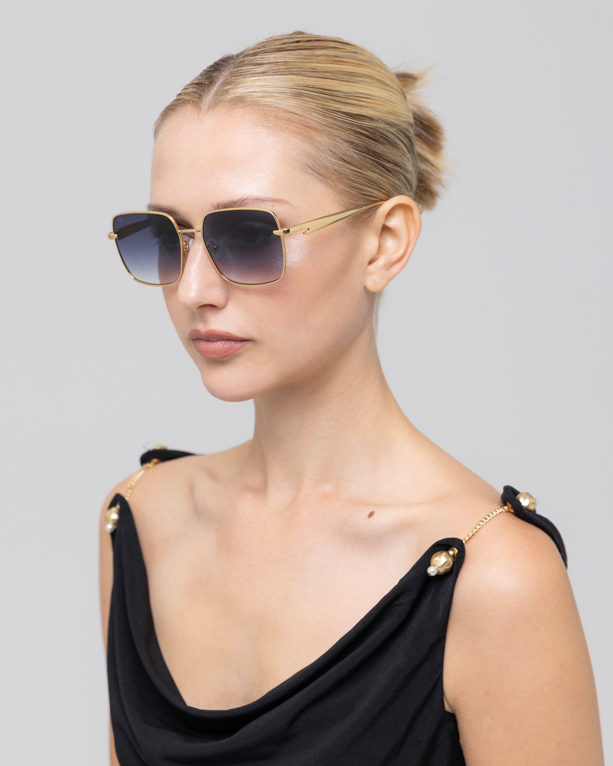 A woman with blonde hair in a neat bun wears For Art's Sake® Edge gold-framed rectangular sunglasses and a black sleeveless top with gold shoulder accents, posing against a plain light gray background.