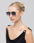 A woman with blonde hair in a neat bun wears For Art's Sake® Edge gold-framed rectangular sunglasses and a black sleeveless top with gold shoulder accents, posing against a plain light gray background.