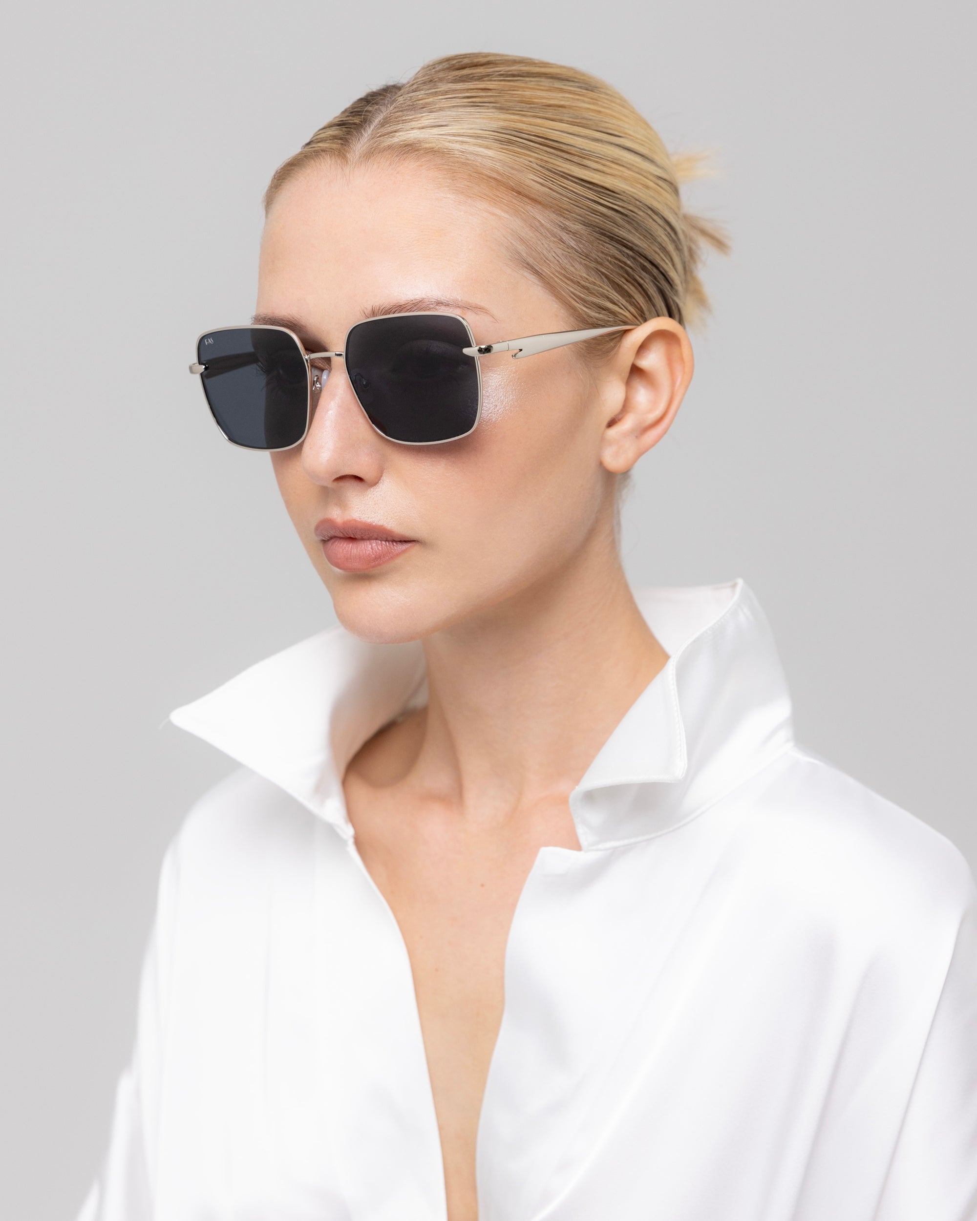 A blonde woman with a low bun wears For Art's Sake® Edge black rectangular sunglasses and a white satin shirt with a sculptural high collar, looking slightly to the side against a plain light background.