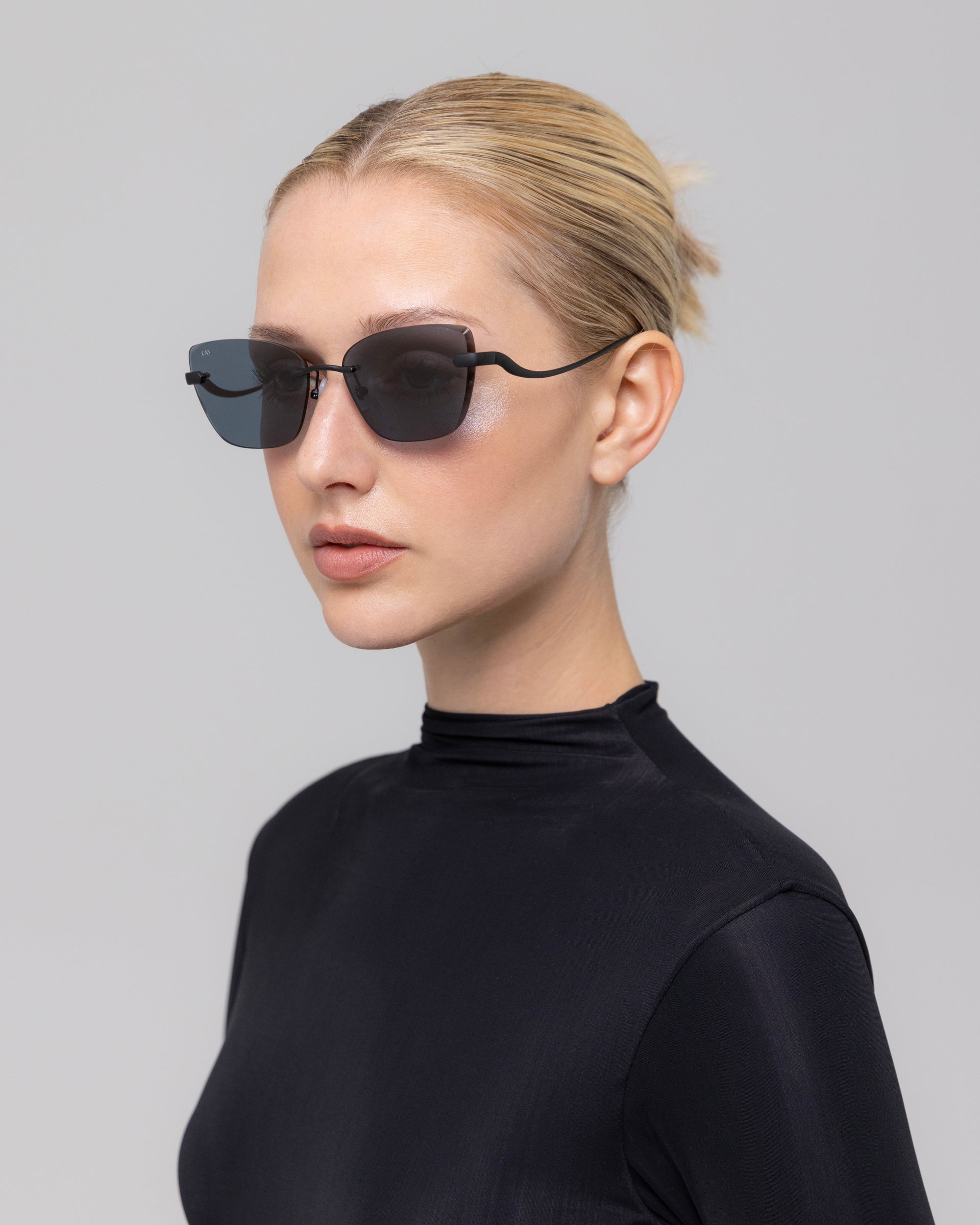 A woman with blonde hair tied back models For Art's Sake® Spark rimless cat-eye sunglasses and a high-neck black top against a plain light gray background.