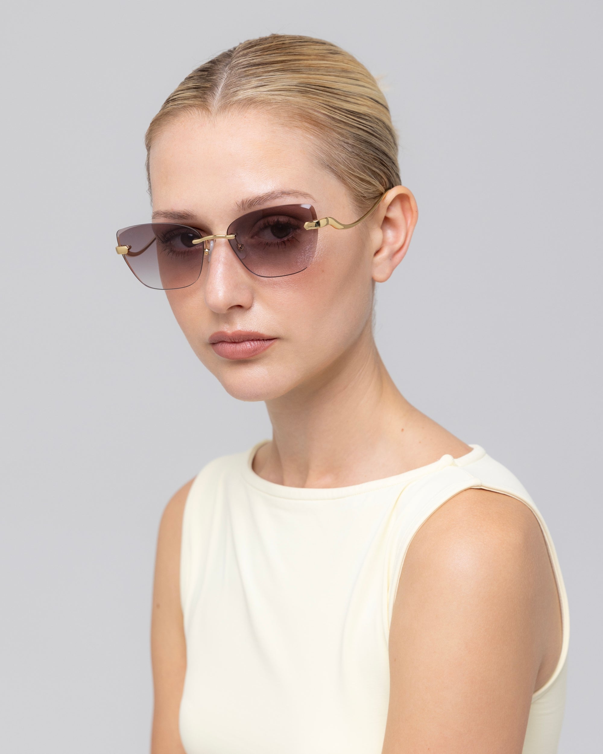 A blonde-haired woman wears For Art's Sake® Spark rimless cat-eye sunglasses with a sleeveless ivory top, posing against a plain light gray background.