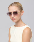 A blonde-haired woman wears For Art's Sake® Spark rimless cat-eye sunglasses with a sleeveless ivory top, posing against a plain light gray background.