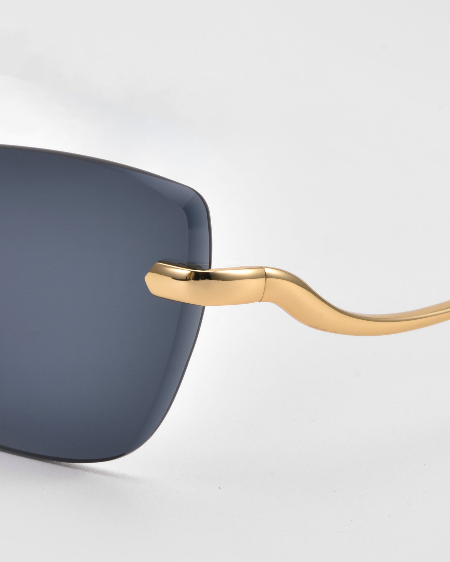 Close-up of For Art's Sake® Spark rimless cat-eye sunglasses with a gold frame and dark tinted lens, highlighting the smooth, curved temple design against a light gray background.