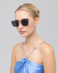 A woman with blonde hair in a neat bun wears For Art's Sake® Spark sculptural sunglasses and a shiny light blue spaghetti strap dress with a chest flower detail, posing against a plain gray background.