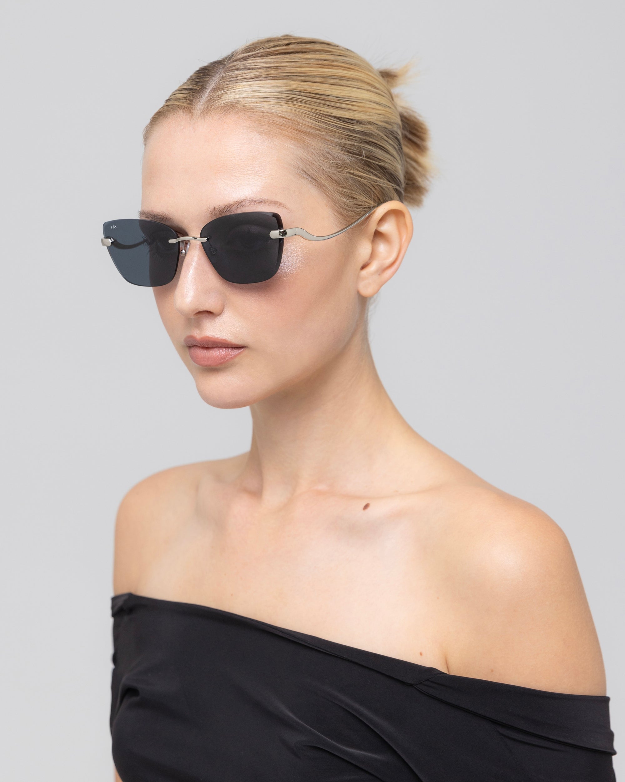 A woman with blonde hair in a neat bun wears For Art's Sake® Spark rimless cat-eye sunglasses and an off-the-shoulder black top, standing against a plain light gray background.