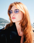 A woman with long red hair wears For Art's Sake® Rebirth sunglasses, showcasing their slim oval silhouette and a black jacket, gazing seriously into the distance with mountains and cityscape in the background under a bright sky.